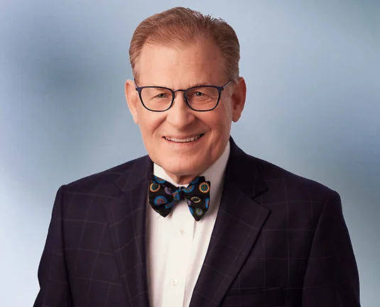 Older Attorney, Fred Dilley, wearing suit with bow tie and square rimmed glasses smiles at camera in front of blue gray background