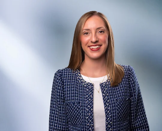 Young woman, Allison Morris, wearing blue checkered blazer white undershirt, smiles at camera in front of blurred blue gray background