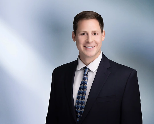 A man, Michael Cassar, with short, dark brown hair and a beard, wearing a black blazer and white shirt, is smiling at the camera, standing against a blurred blue and white background.”