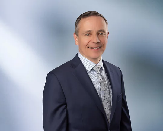 “A man, Matt Hrebec, with short, light brown hair, wearing a grey blazer and white collared shirt, is smiling at the camera, standing against a blurred blue and white background.”