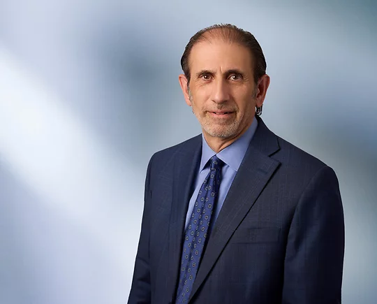 A man, Jonathan David, in a dark blue suit, light blue shirt, and patterned tie is looking at the camera, standing against a blurred blue and white background.