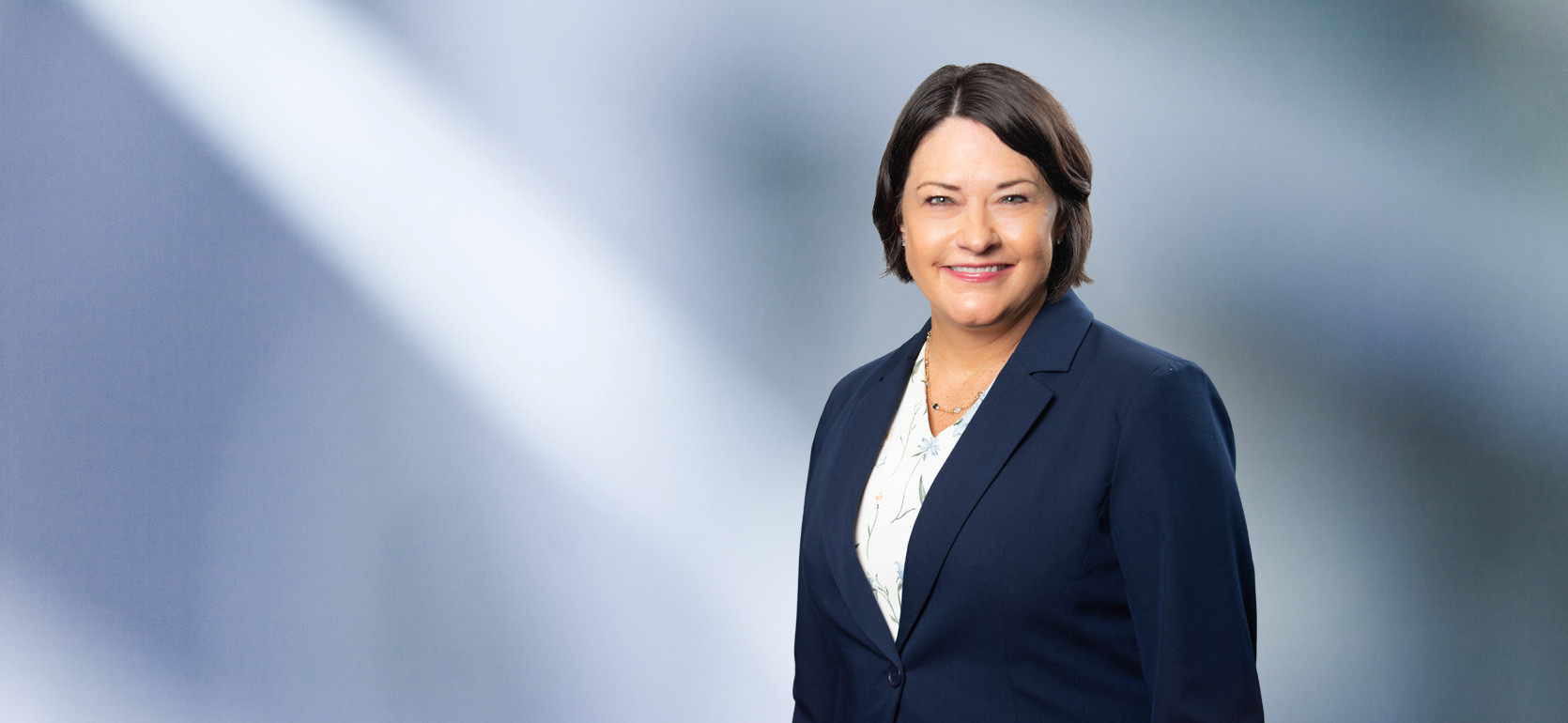 Woman, Marcy Hahn, with short dark hair smiles at camera wearing blue blazer and white floral shirt in front of blurred blue gray background