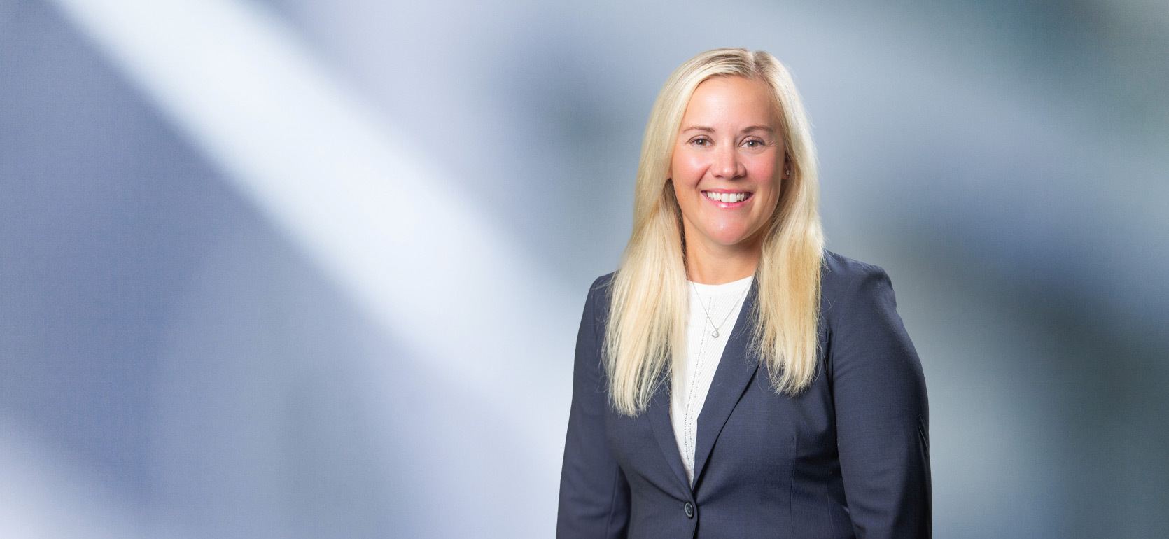 A woman, Lisa Serra, with blond hair and wearing a gray sport coat and white undershirt, smiles at camera in front of blurred blue gray background