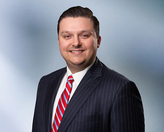 A man, Joe Giacalone, smiles at camera wearing a black pinstripe suit, white shirt and red striped tie in front of blue gray blurred background