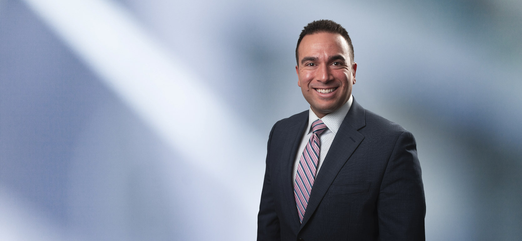 A man, Adam Fadly, in a dark suit, light shirt, and striped tie is smiling at the camera, standing against a blurred blue and white background.