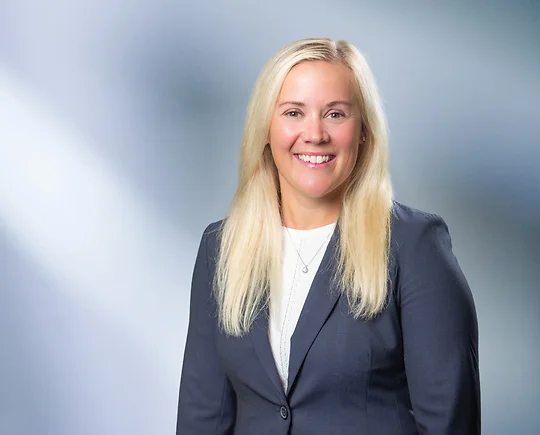 A woman, Lisa Serra, with blond hair and wearing a gray sport coat and white undershirt, smiles at camera in front of blurred blue gray background