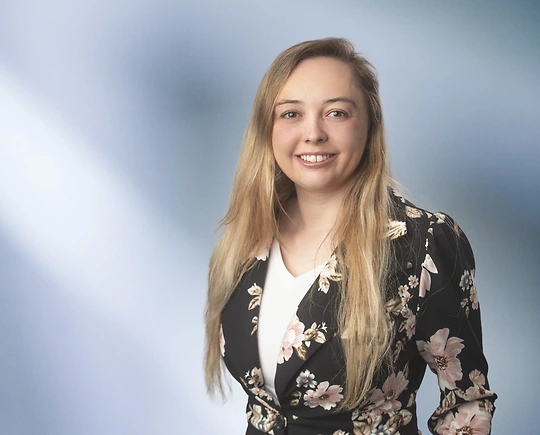 A woman, Jordan Asbury, in a black blazer with a white blouse and a floral pattern is smiling at the camera, standing against a blurred blue and white background.
