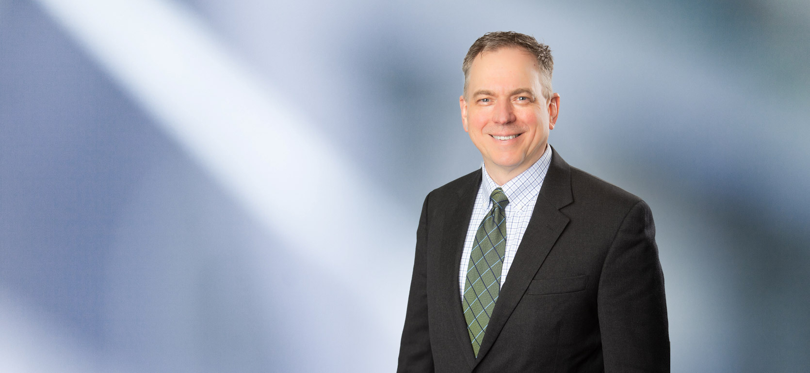 A middle-aged man, Thomas Huyck, smiles at camera, wearking a black sport coat with stripped patterned collared shirt and green necktie in front of blue gray blurred background