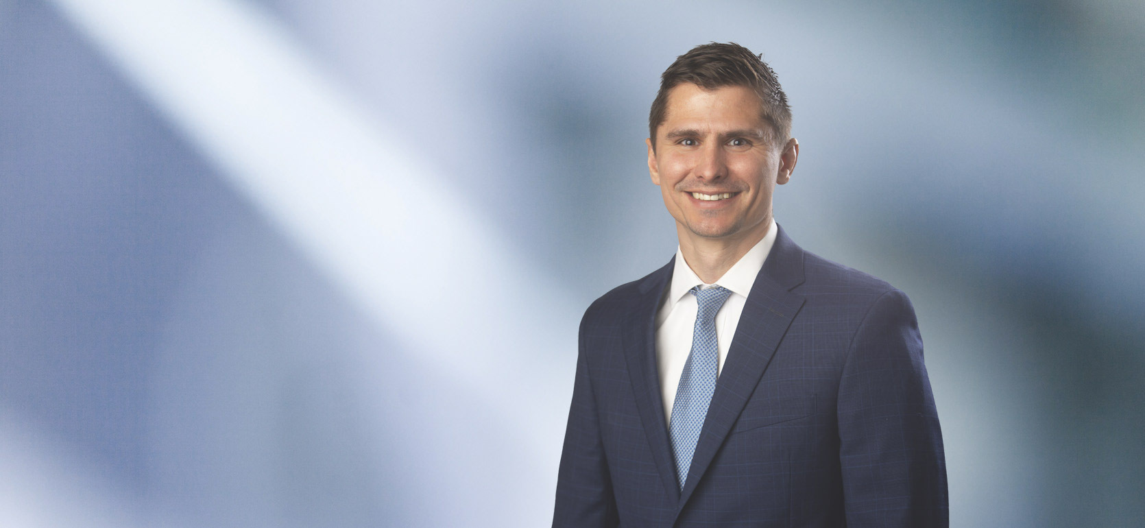 Man, Nicholas Oertel, with light brown hair wearing a charcoal blazer, smiling against a blurred white and blue background.