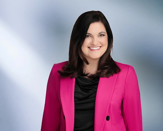 Woman, Stefania Gismondi, wearing a pink blazer, standing against a blurred blue and white background.