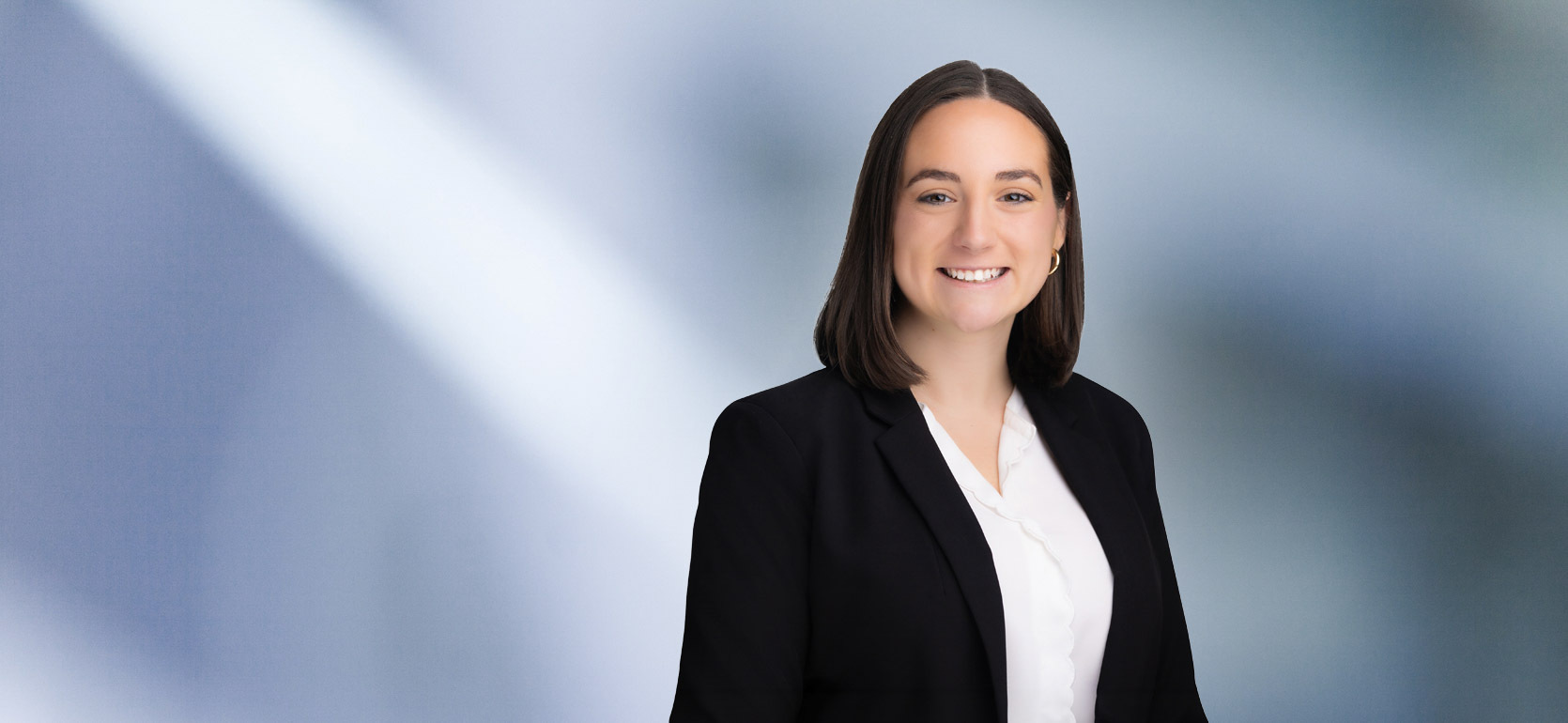 woman, Samantha Shepard, with dark brown hair, wearing a black blazer, smiling against a blurred blue and white background"