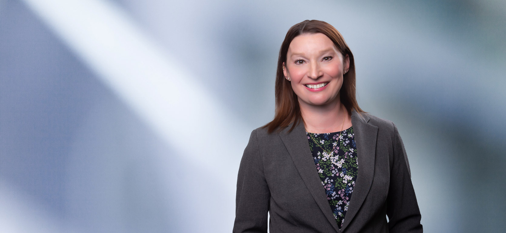 Young woman with brown hair, Laura Kane, smiles at camera wearing gray blazer and flower pattern black undershirt in front of blurred blue gray backround