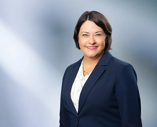 Woman, Marcy Hahn, with short dark hair smiles at camera wearing blue blazer and white floral shirt in front of blurred blue gray background