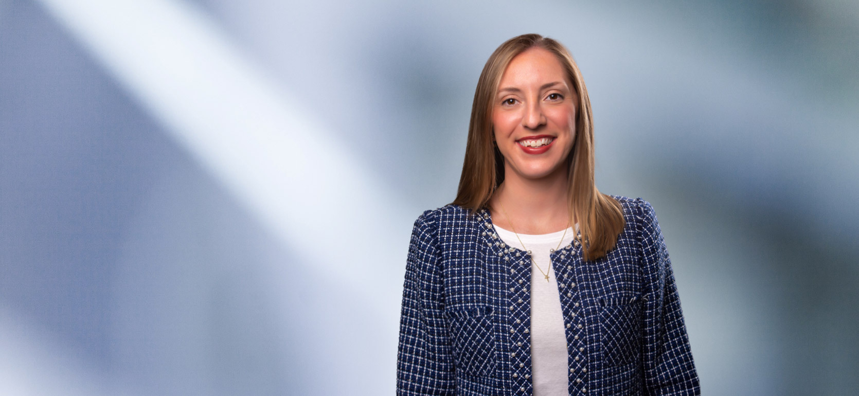 Young woman, Allison Morris, wearing blue checkered blazer white undershirt, smiles at camera in front of blurred blue gray background