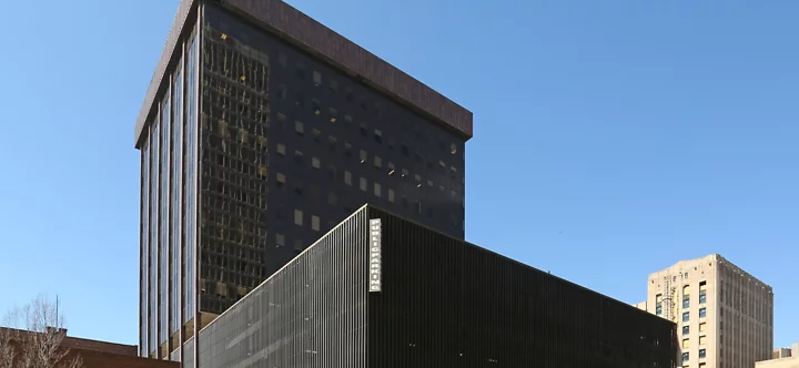 Exterior view of a tall, dark-glass skyscraper in downtown Detroit with a modern facade. The building reflects nearby structures in its windows. A connected lower structure with vertical black slats features a “Public Parking” sign.