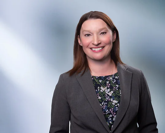 Young woman with brown hair, Laura Kane, smiles at camera wearing gray blazer and flower pattern black undershirt in front of blurred blue gray backround