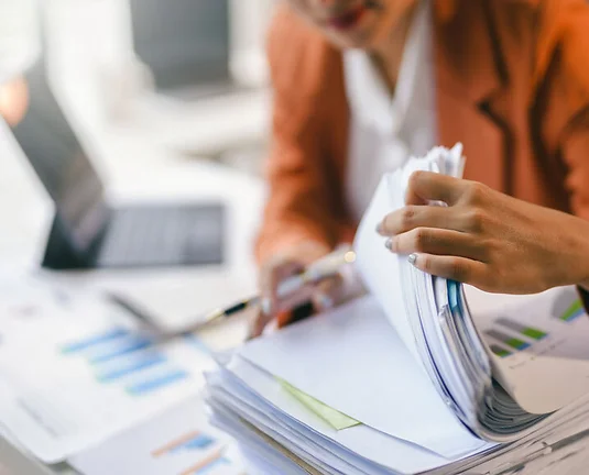 Businesswoman is searching through a stack of paperwork at her desk, looking for financial information