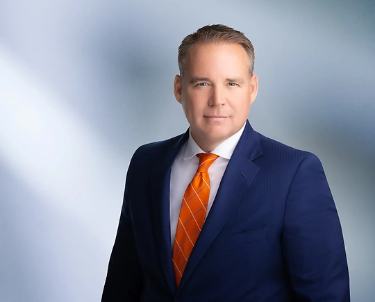 A man, Davis Russell, in a navy blue suit, white shirt, and orange patterned tie is looking at the camera, standing against a blurred blue and white background.