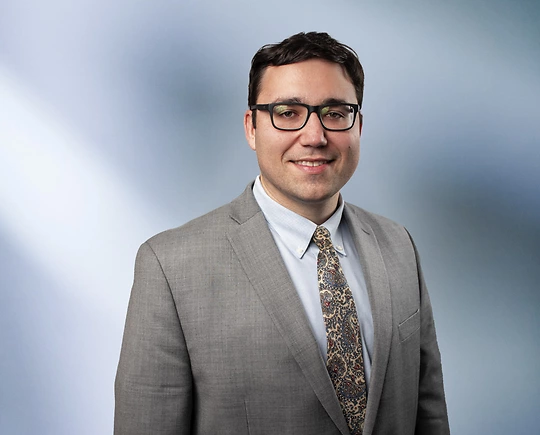 A man, Jeffrey Schultz, in a light gray suit, pale blue shirt, and paisley tie is smiling at the camera, standing against a blurred blue and white background.