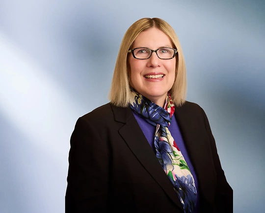 A woman, Jennifer Vanregemorter, in a black blazer, purple top, and multicolored floral scarf is smiling at the camera, standing against a blurred blue and white background.