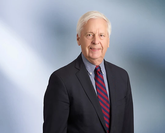 A man, Lynwood Vandenbosch, in a dark suit, checkered shirt, and red and blue striped tie is smiling at the camera, standing against a blurred blue and white background.