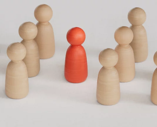 Group of light wooden figurines surrounding a single red figurine, symbolizing individuality or leadership.