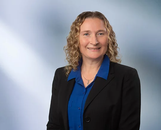 Woman with curly blonde shoulder length hair, Jennifer Knox, wearing black blazer and dark blue collared button-down shirt, smiles at camera in front of blue gray background