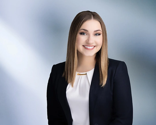 A woman, Lindsey Mead, with straight, light brown hair, wearing a navy blazer and white top with a gold accent, is smiling at the camera, standing against a blurred blue and white background.