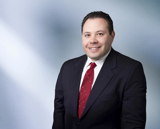 Smiling man, Ben Price, wearing a dark suit with a white shirt and red patterned tie, standing in front of a soft blue and white blurred background
