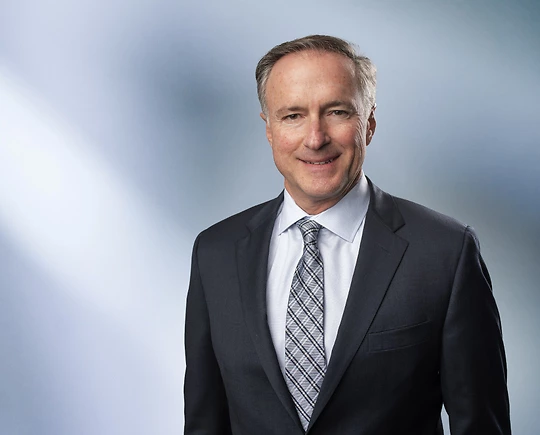 A man, Paul Millenbach, in a dark suit, light shirt, and patterned tie is smiling at the camera, standing against a blurred blue and white background.