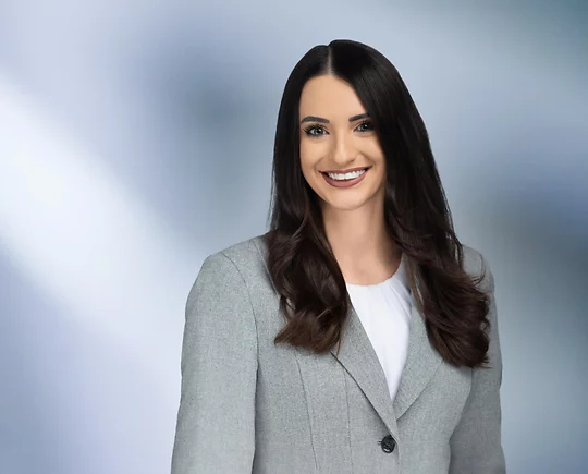 “A woman, Melanie Assad, with long, dark brown hair, wearing a grey blazer and white top, is smiling at the camera, standing against a blurred blue and white background.”
