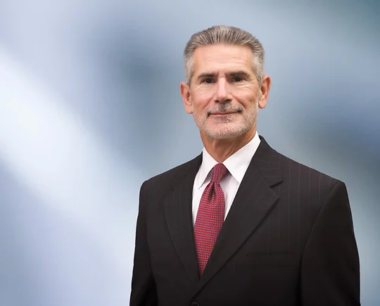 A man, Lawrence Korolewicz, in a dark pinstripe suit, white shirt, and red patterned tie is looking at the camera, standing against a blurred blue and white background.