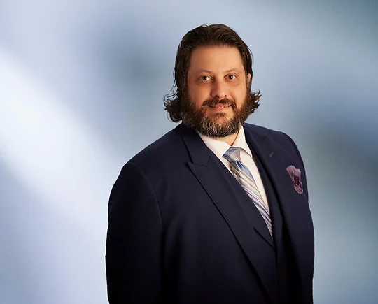 Smiling man, Robert Hamor, with medium-length dark hair and a beard, wearing a navy blue suit, striped tie, and pocket square, standing in front of a soft blue gradient background.