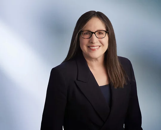 Smiling woman, Julie Fershtman, with long straight brown hair and glasses, wearing a dark blazer, standing in front of a soft blue gradient background