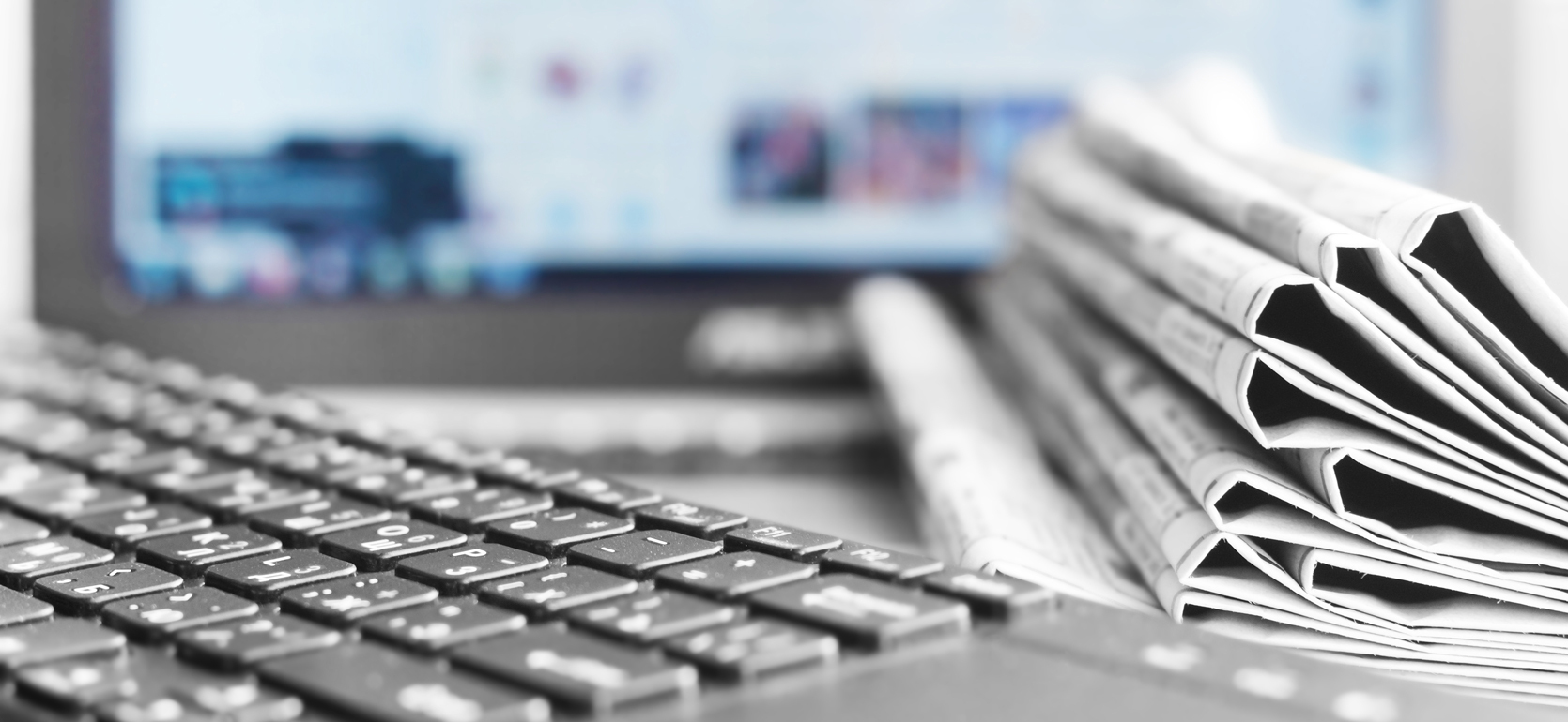 Keyboard next to stack of newspapers in front of blurred computer screen.