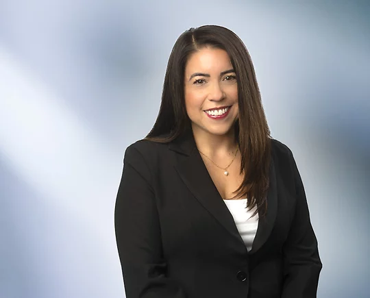 An attractive woman, Jacquelyn Dupler, in a white blouse with a black blazer is smiling at the camera, standing against a blurred blue and white background.