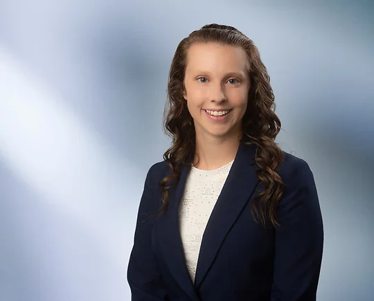A young woman, Allison Shudark, wearing a black sport coat and white lace blouse, smiles at camera in front of blurred blue-white background