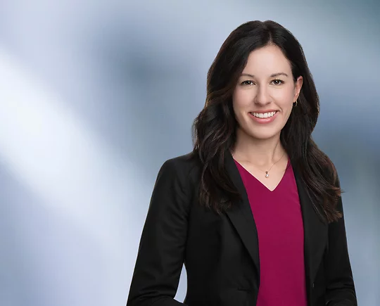 A woman, Julie Hamlet, in a black blazer and purple top is smiling at the camera, standing against a blurred blue and white background.