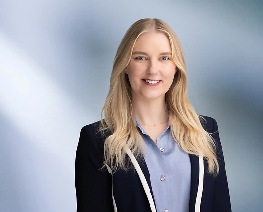 A woman, Alaina Nelson, with long blonde hair wearing a navy blazer over a light blue blouse stands in front of a blurred, blue-toned background.