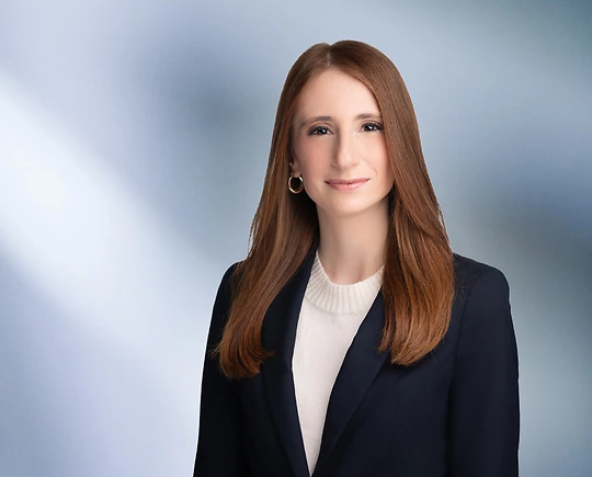Smiling woman, Dina Kashat, with long, straight auburn hair, wearing a dark blazer over a white top, standing against a soft blue gradient background