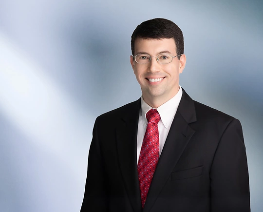 A man, James Anderton, in a black suit, white shirt, and red patterned tie is smiling at the camera, standing against a blurred blue and white background.