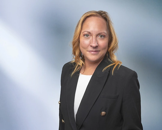 A woman, Elizabeth Cotner, in a black blazer with gold buttons and a white top is looking at the camera, standing against a blurred blue and white background.