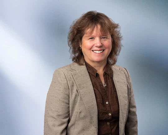 A woman, Kimberly Hafley, in a tan blazer and dark brown shirt is smiling at the camera, standing against a blurred blue and white background.