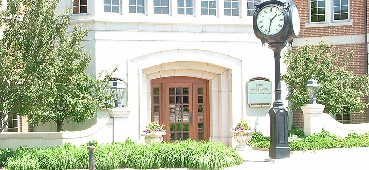 Front entrance of the Holland, Michigan office with arched stone doorway, large wooden double doors, and a classic black street clock. Surrounded by greenery, flowers, and brick architecture with a visible office sign reading “Baird Bayside Capital.”
