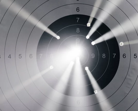 A paper shooting target with several bullet holes clustered near the center, illuminated by dramatic beams of light converging toward the bullseye.