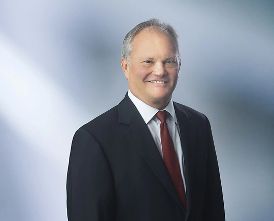 A man, Brian Goodenough, in a dark suit, white shirt, and red tie is smiling at the camera, standing against a blurred blue and white background.