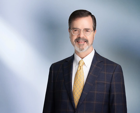 A man, Jeffrey Theuer, in a dark checkered suit, white shirt, and yellow patterned tie is smiling at the camera, standing against a blurred blue and white background.