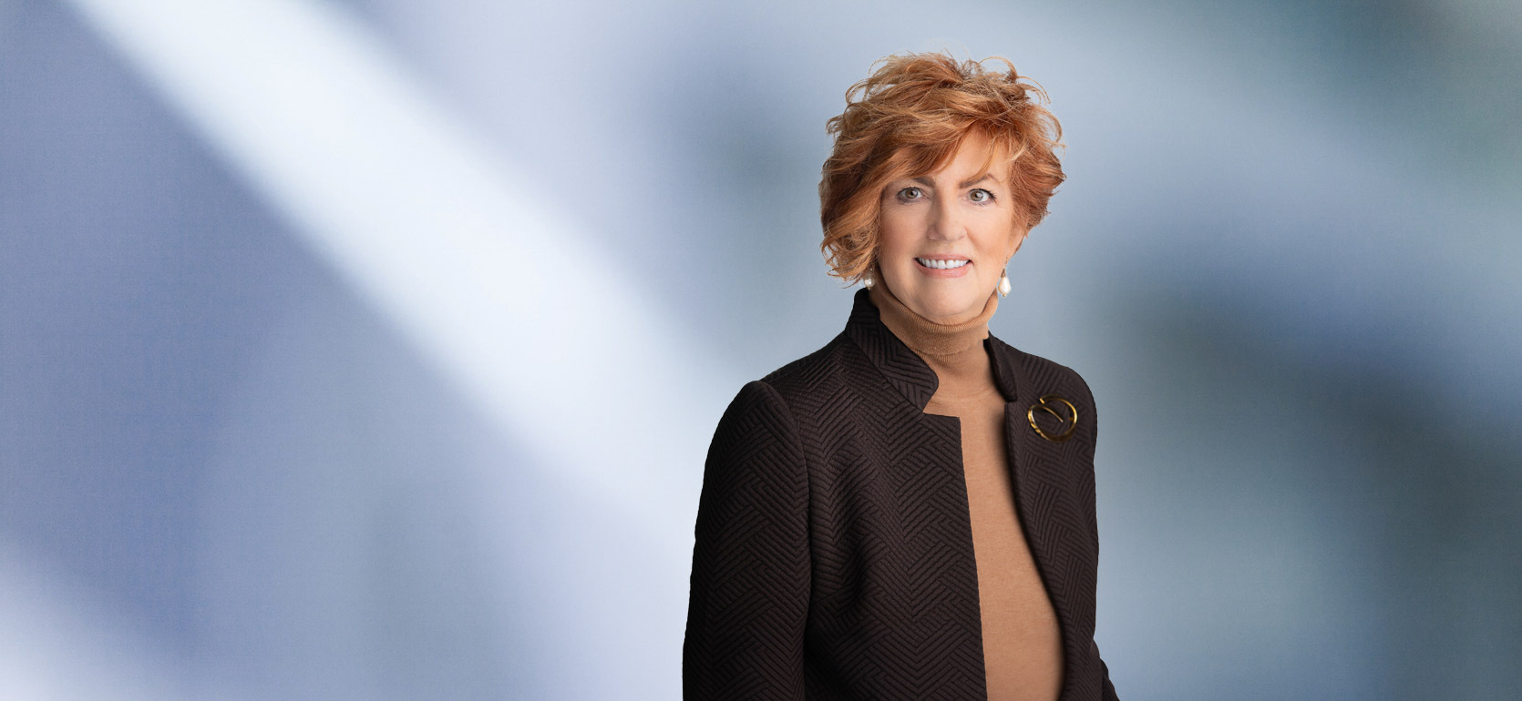 A woman, Kelly Lucas, in a brown textured blazer over a tan turtleneck, accessorized with pearl earrings and a brooch, is smiling at the camera, standing against a blurred blue and white background.