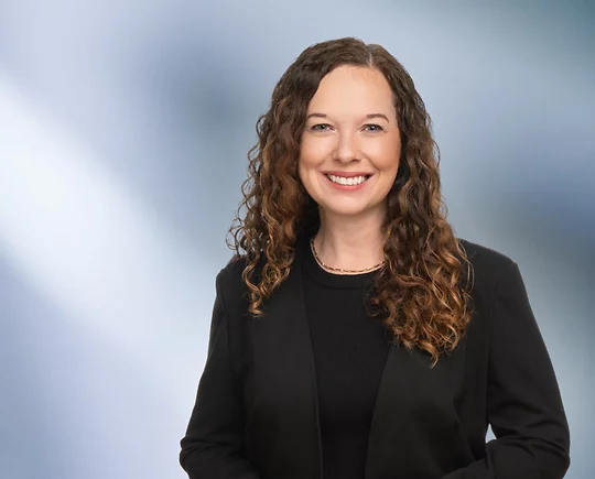 A woman, Laura Genovich, with curly brown hair, wearing a black blazer and black top, is smiling at the camera, standing against a blurred blue and white background.