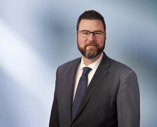 “A man, Mark Koerner, with short, dark brown hair and glasses, wearing a grey blazer and white shirt, is smiling at the camera, standing against a blurred blue and white background.”